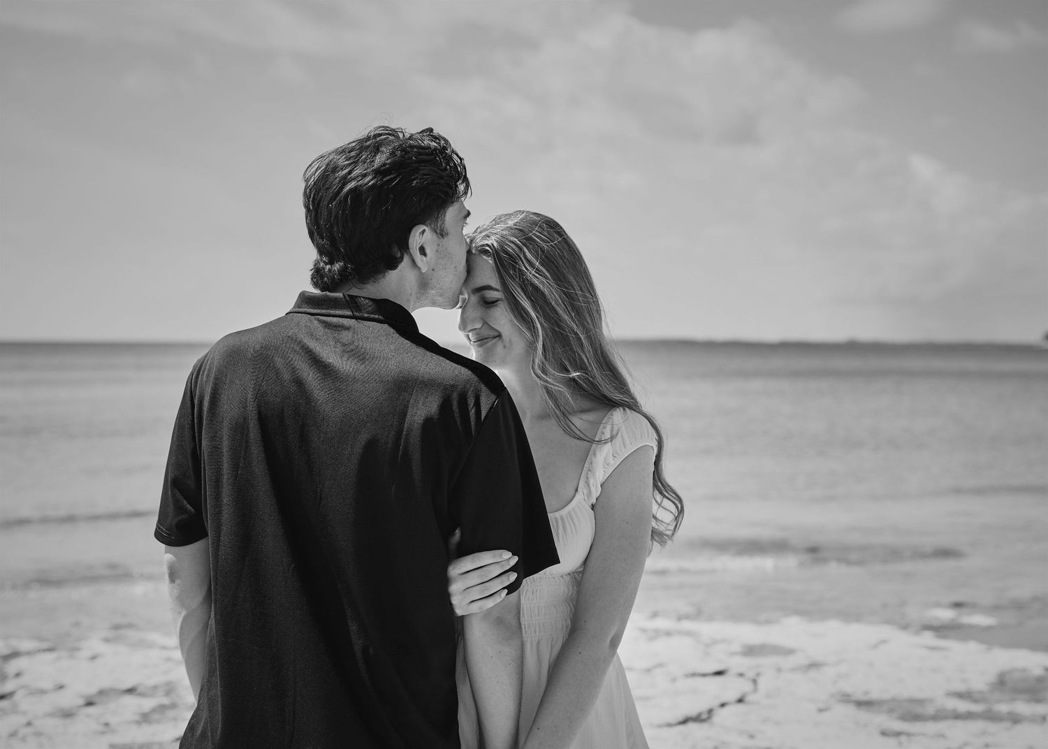 Black and white photo of a romantic couple sharing a tender moment on a beach with ocean in the background.
