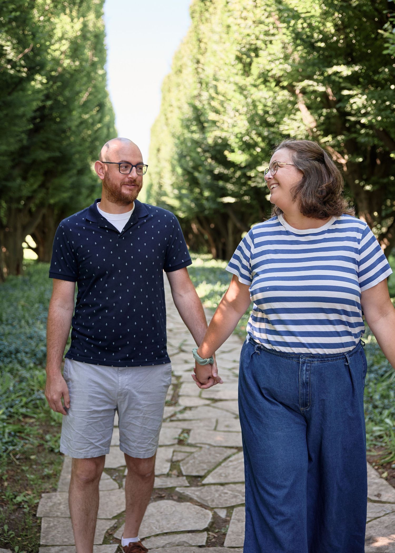 Couple holding hands and smiling at each other on a stone path lined with tall green trees.