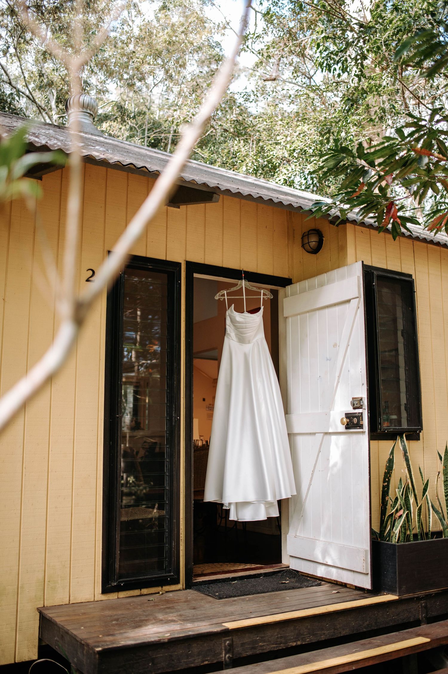 A white wedding dress hangs outside a yellow cabin with rustic wooden details.