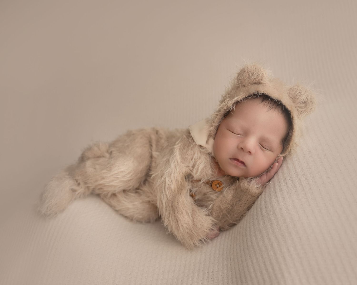 A sleeping infant wearing a furry beige outfit with bear ears lies on a textured cream backdrop.