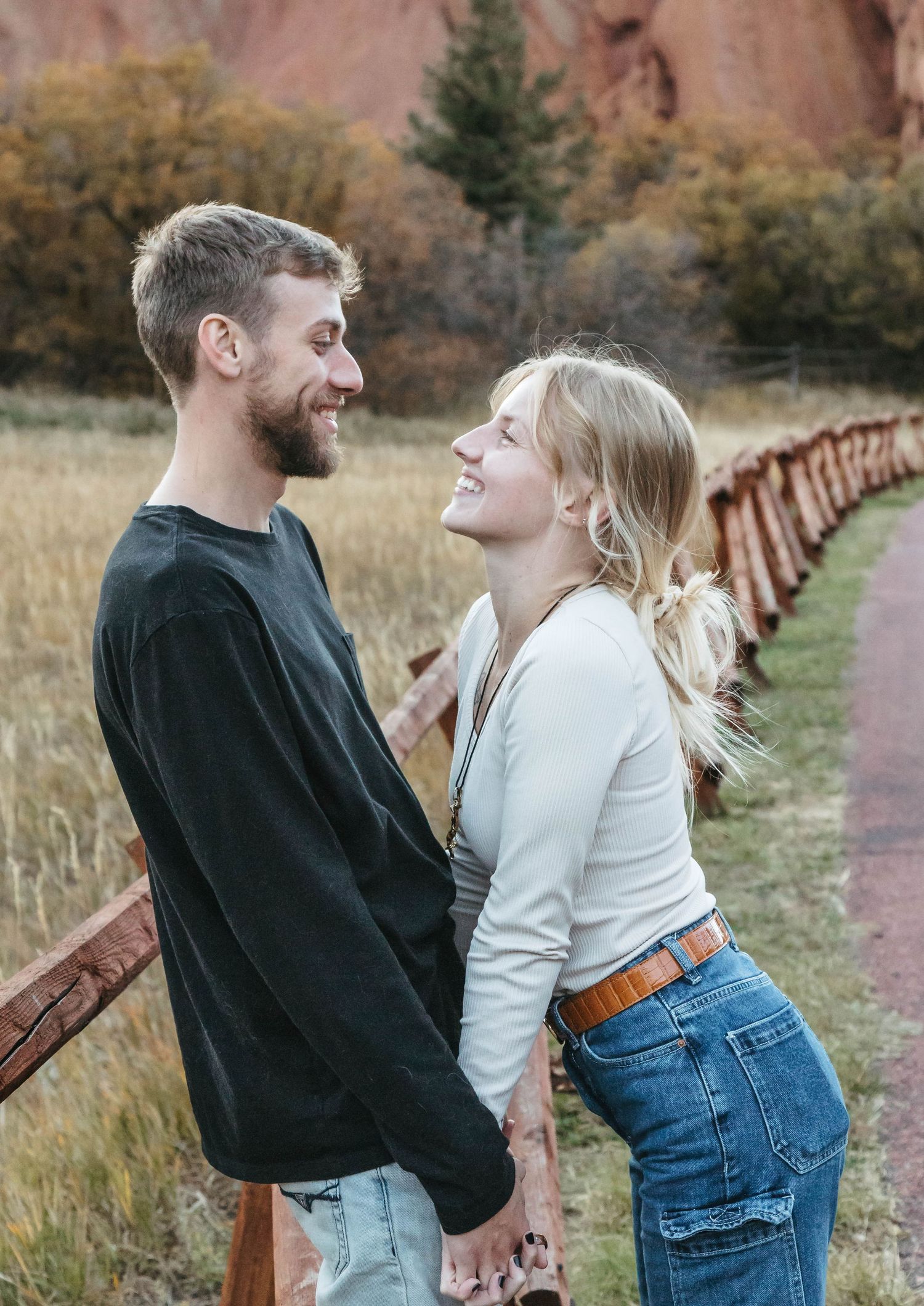 A couple shares a joyful moment together on a path with rustic wooden fencing and red rock formations in the background.