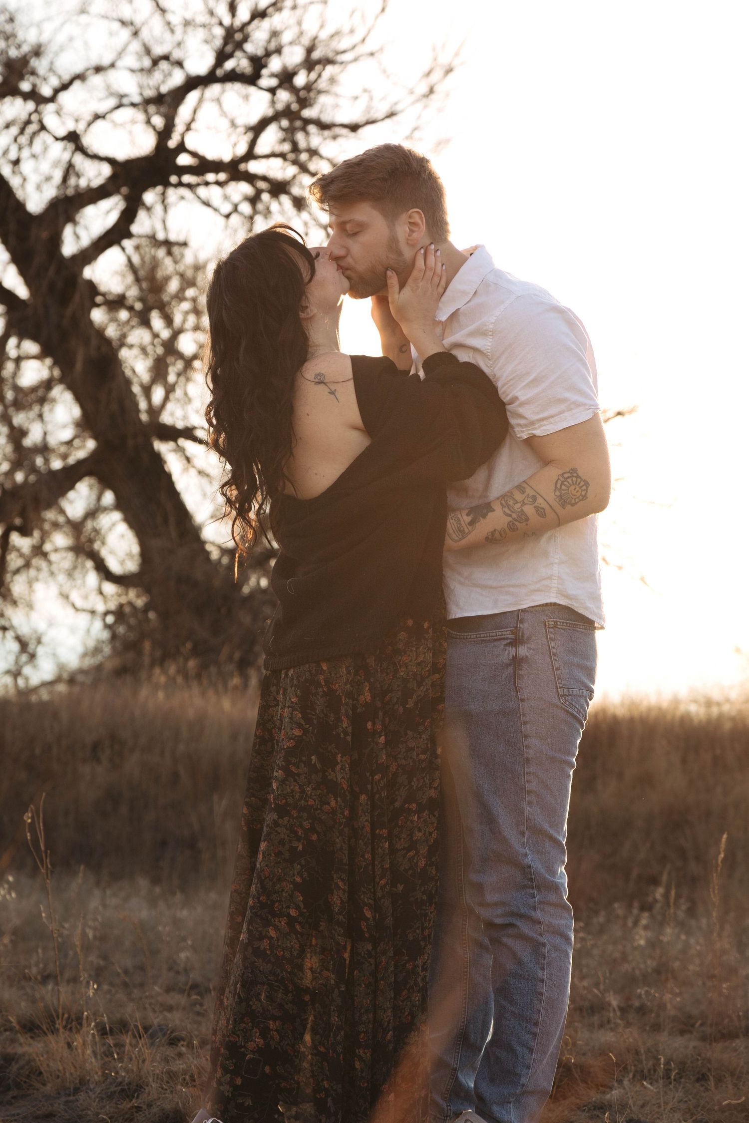 A romantic couple embraces in a field at sunset with bare trees silhouetted in the background.