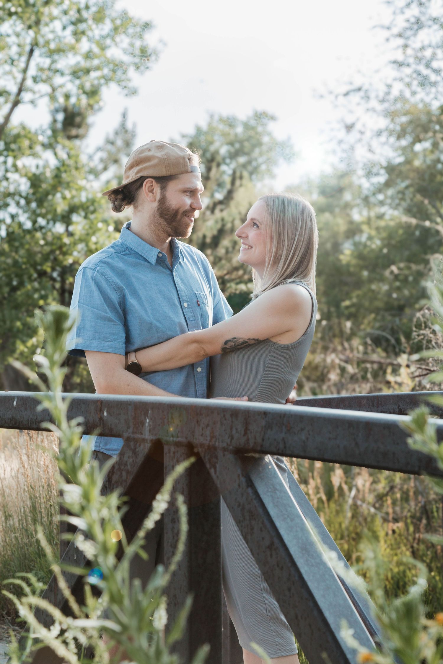 A couple gazes at each other while standing on a wooden bridge surrounded by greenery.