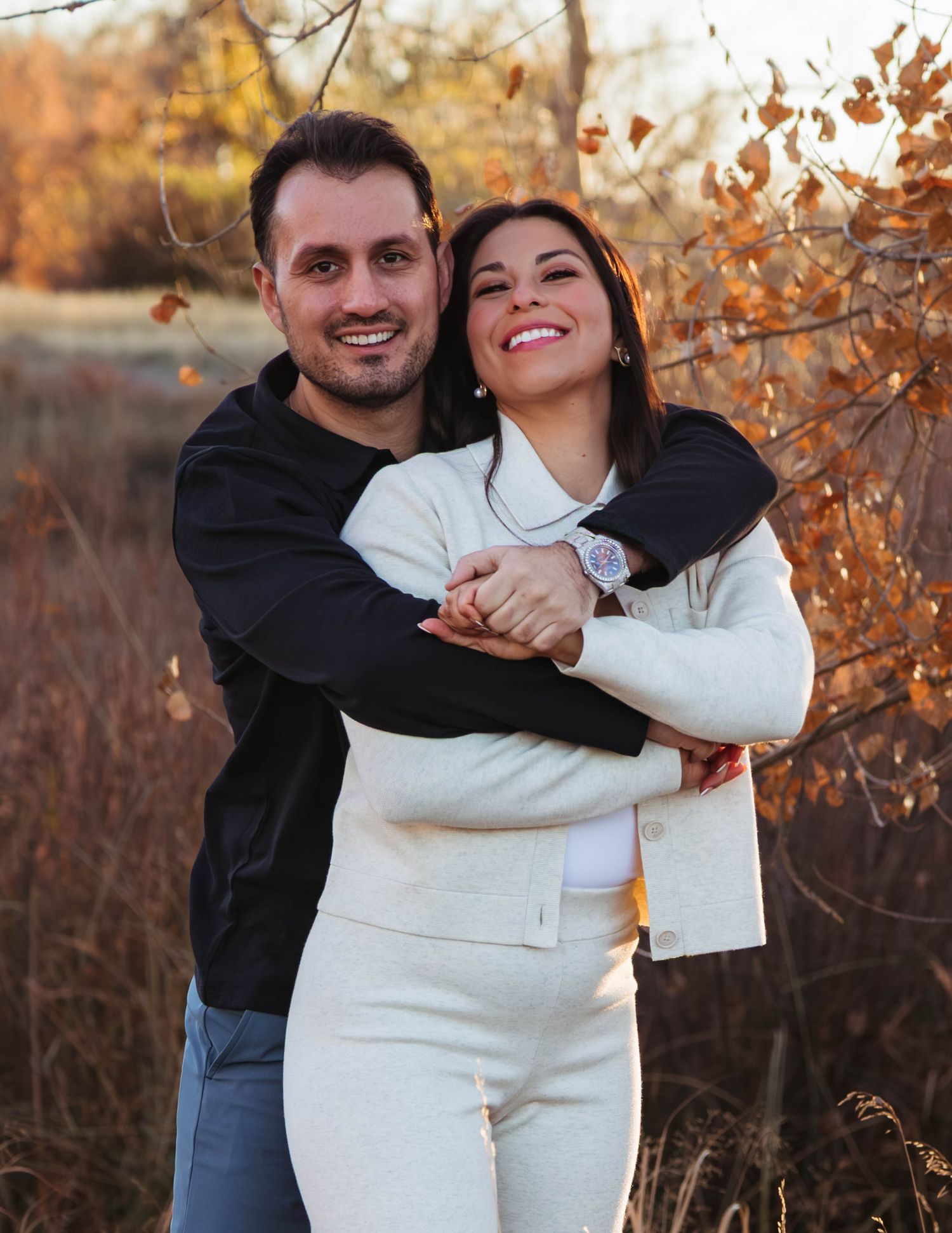 A couple embraces and laughs together in a natural setting with autumn foliage during golden hour.