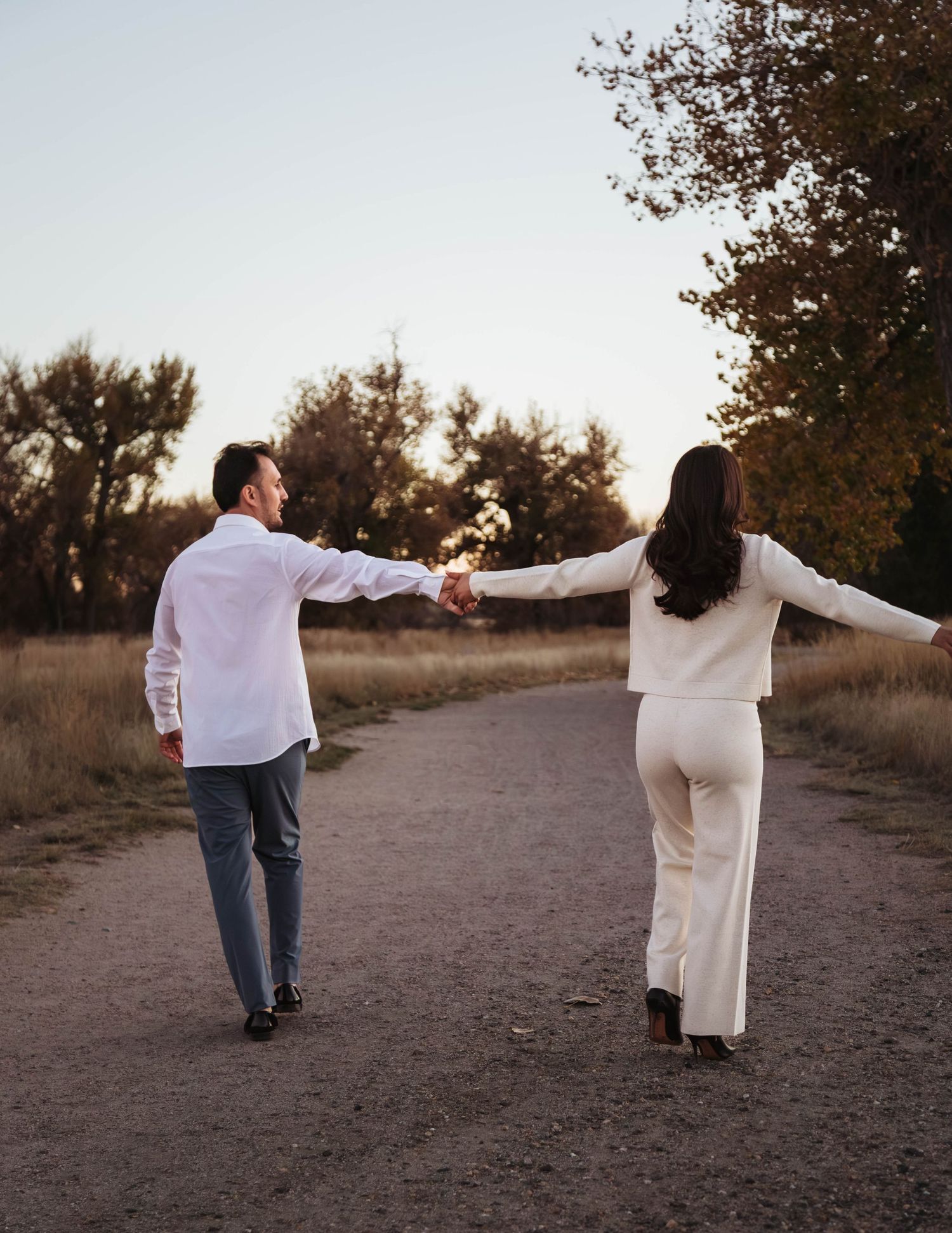 Couple walking together on a dirt path at sunset while holding hands and walking apart in a playful manner.