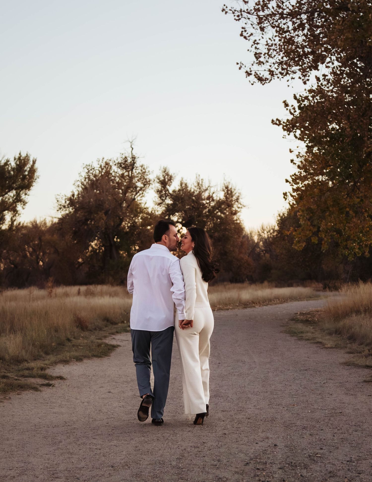 A couple walking hand in hand on a dirt path at sunset with autumn trees and golden light in the background.