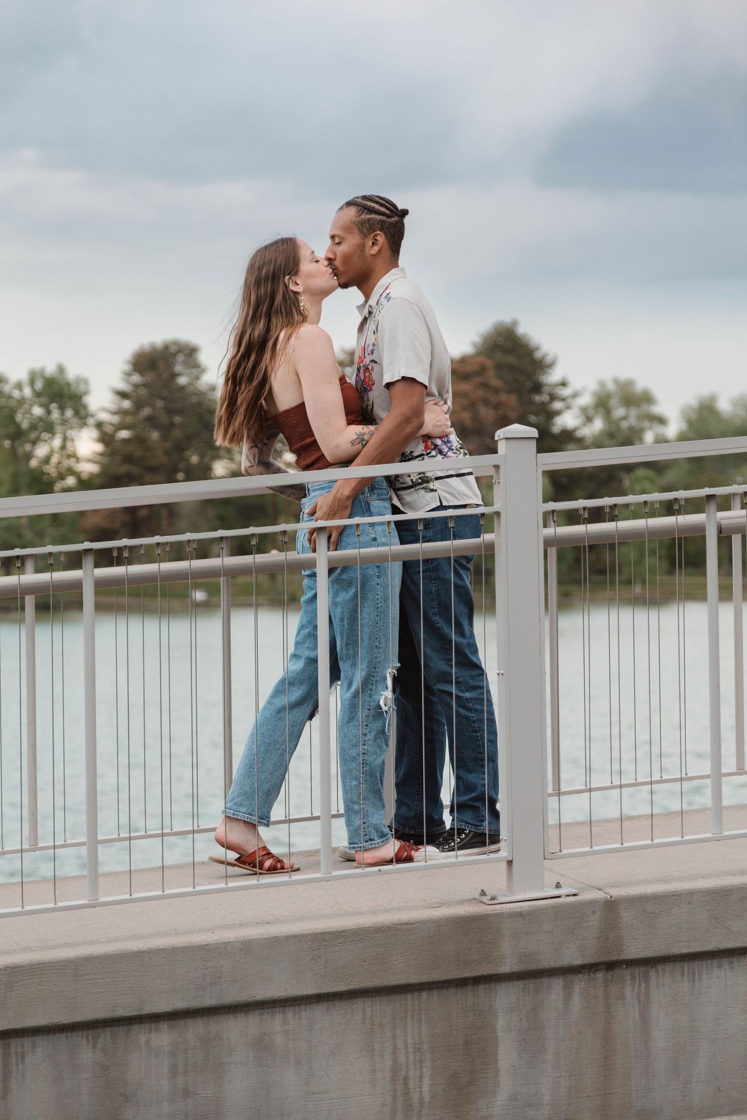 Couple embracing on a bridge railing over water during sunset.