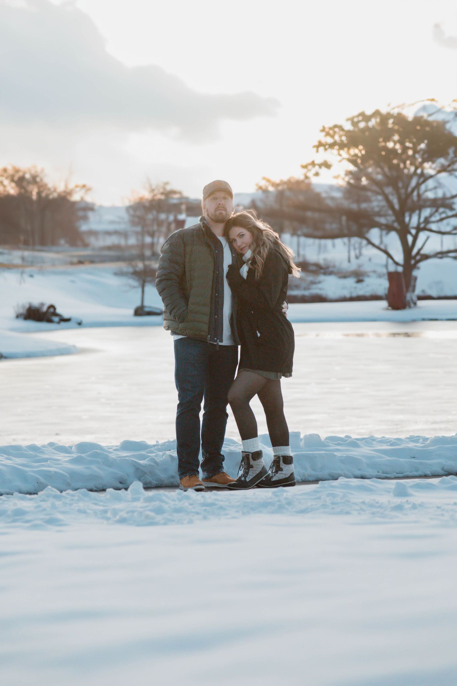 A romantic winter photoshoot of a couple standing together in the snow at sunset.