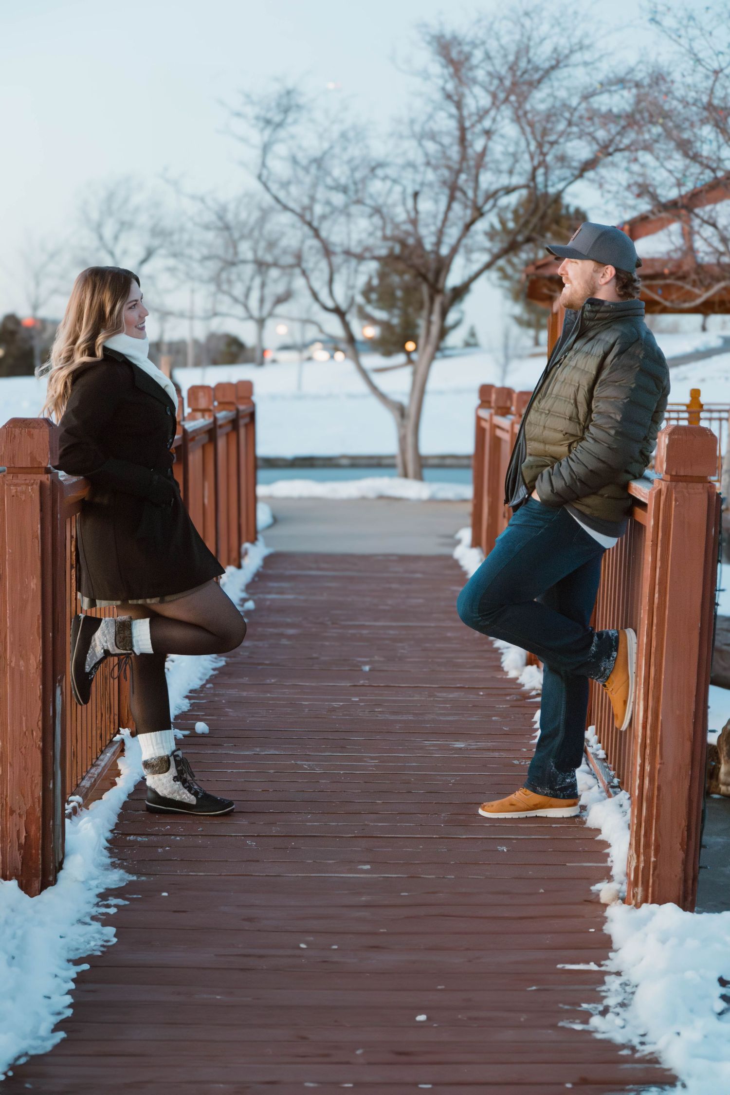 A playful winter photo of a couple leaning on opposite sides of a wooden bridge railing.