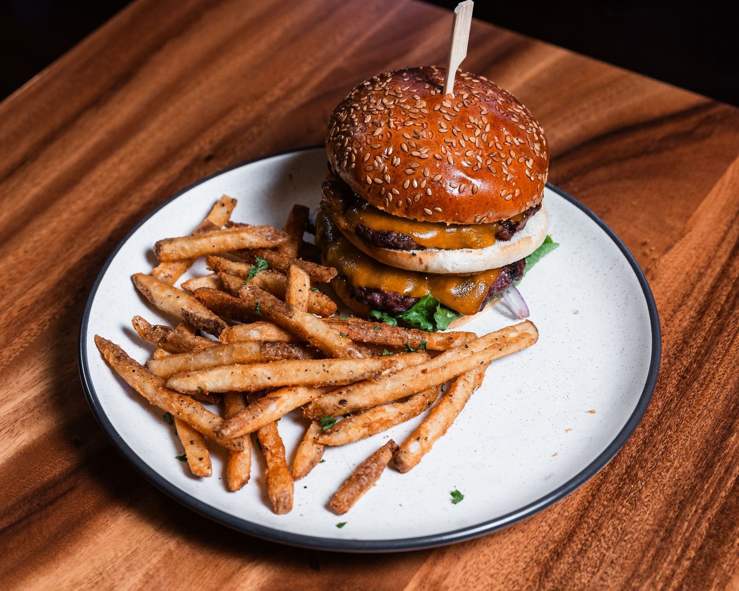 Sesame seed burger with toothpick garnish served alongside seasoned french fries on white plate.