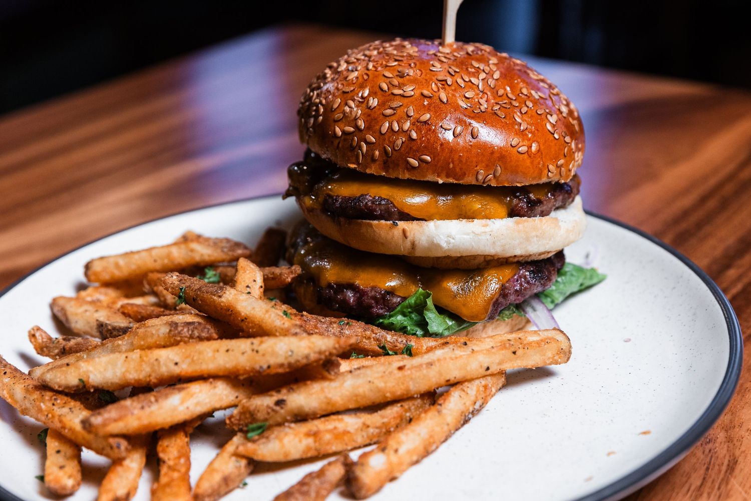 Juicy hamburger with sesame seed bun served alongside crispy french fries on a white plate against wooden surface.