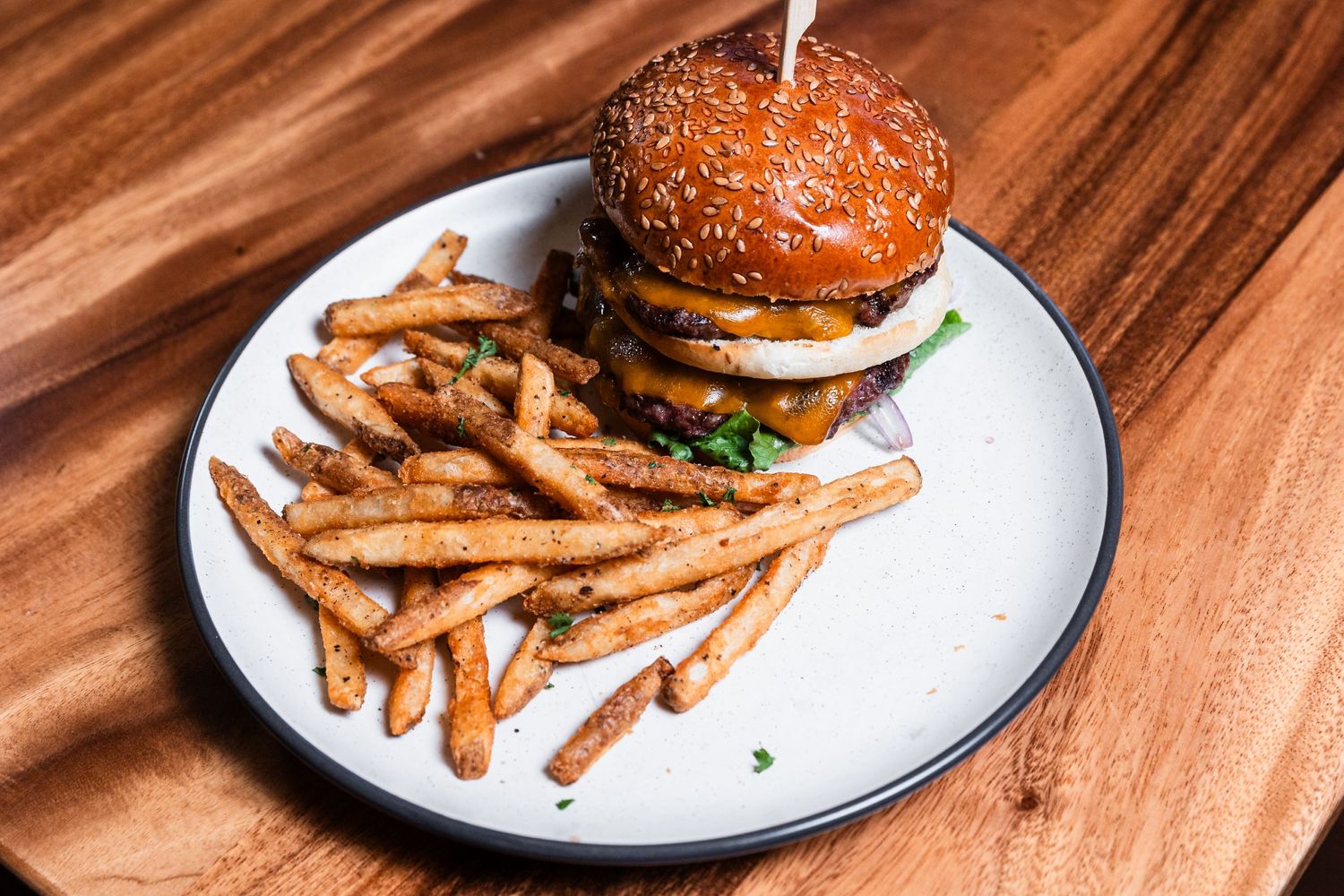 Double-decker burger with sesame seed bun and side of seasoned french fries on ceramic plate.