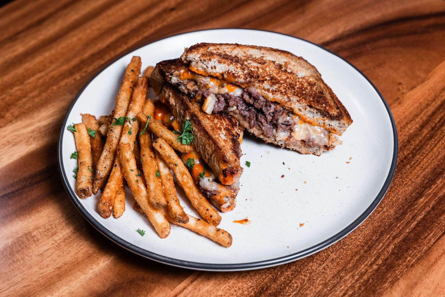 Grilled sandwich cut in half revealing melted filling, served with seasoned french fries on white plate.