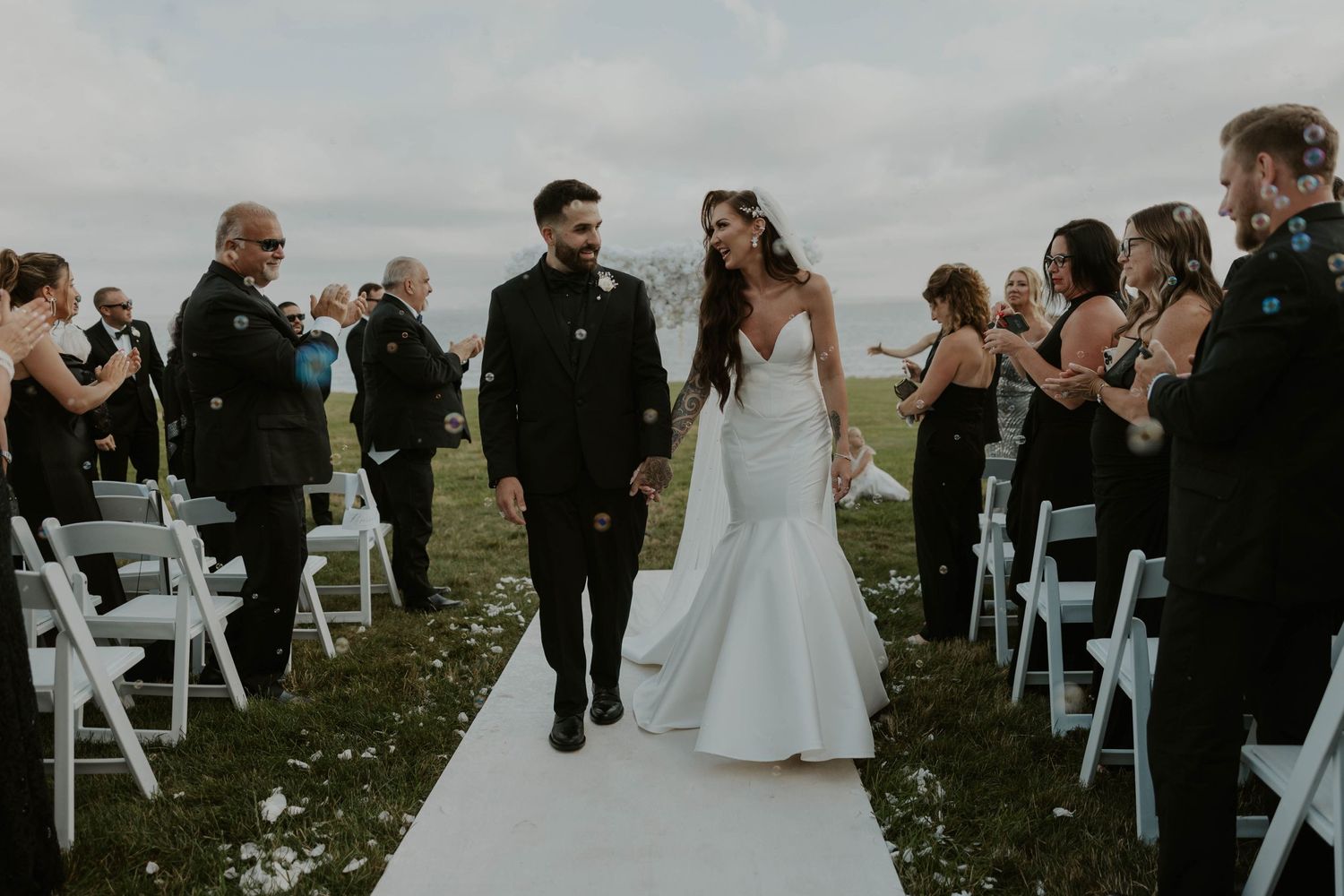 A beautiful outdoor wedding ceremony with guests seated on white chairs along a white aisle runner.