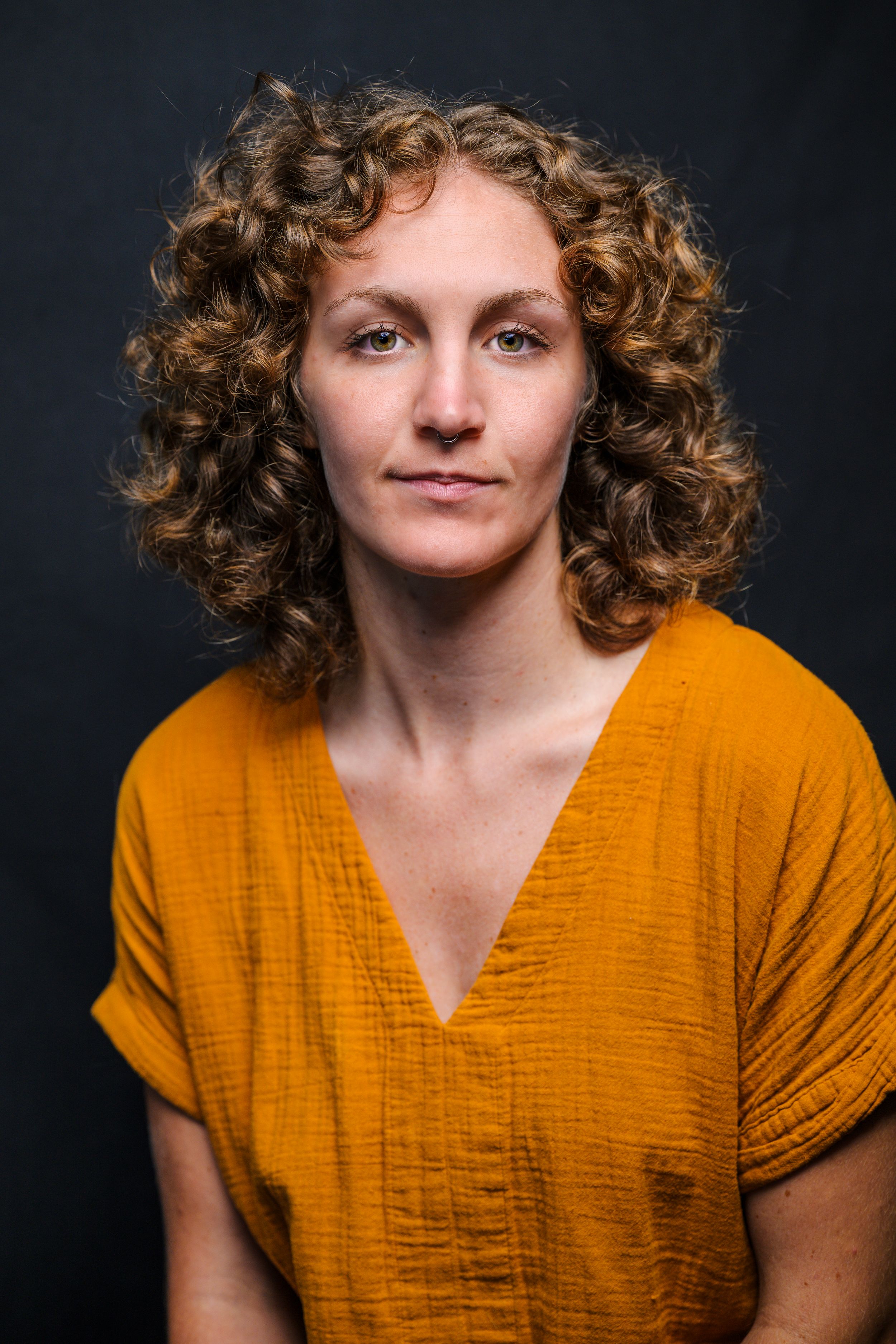 Portrait of a person with curly brown hair wearing a mustard yellow v-neck top against a dark background.