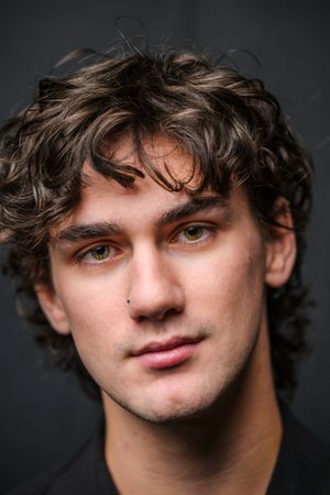 Portrait against dark background showing curly hair, nose piercing and gentle smile in moody lighting.