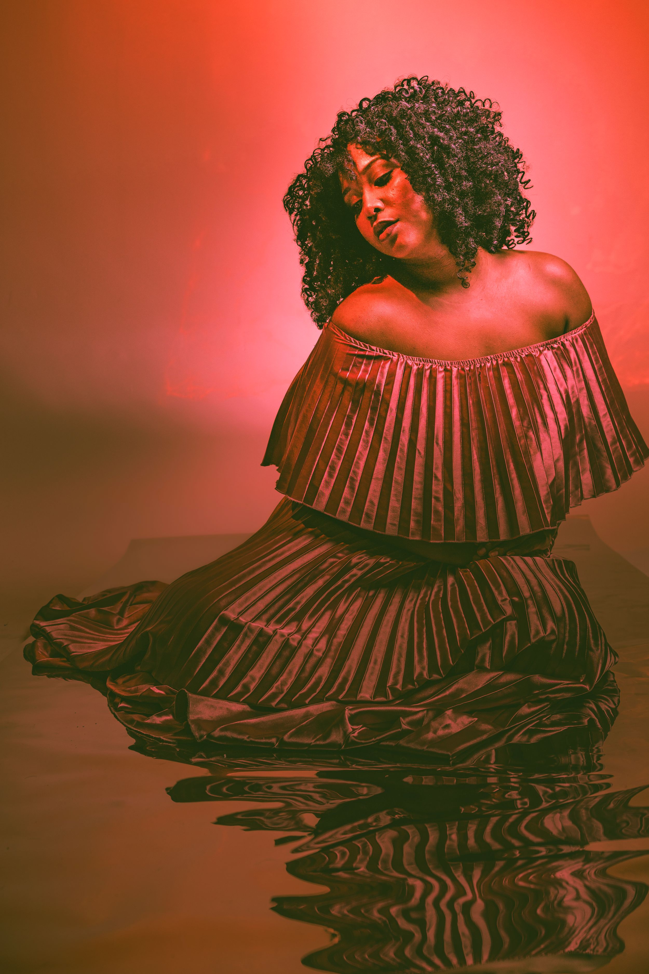 A dramatic fashion portrait in red lighting shows a model in a pleated black dress reclining in an artistic pose.