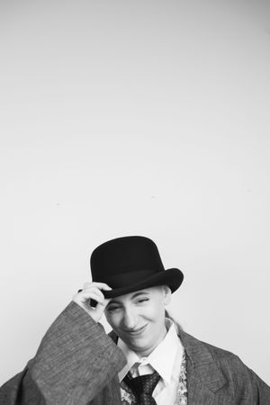 Black and white photo of a person wearing a bowler hat and suit smiling in a playful pose against a light background.