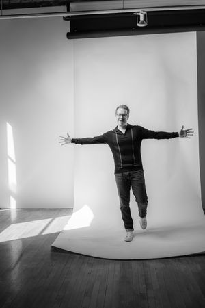 A person in black clothing jumps with arms outstretched against a white studio backdrop in a black and white photo.