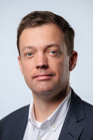 Professional headshot portrait of person in business attire with gray blazer and white button-down shirt against light background.