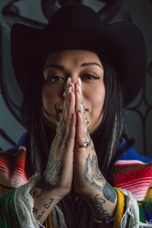 A person with tattooed hands presses their palms together in front of their face while wearing colorful clothing.
