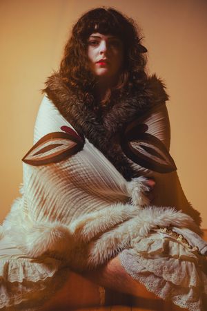 A figure in a dramatic white ruffled dress and dark curly hair poses against a warm-toned background in a vintage style.