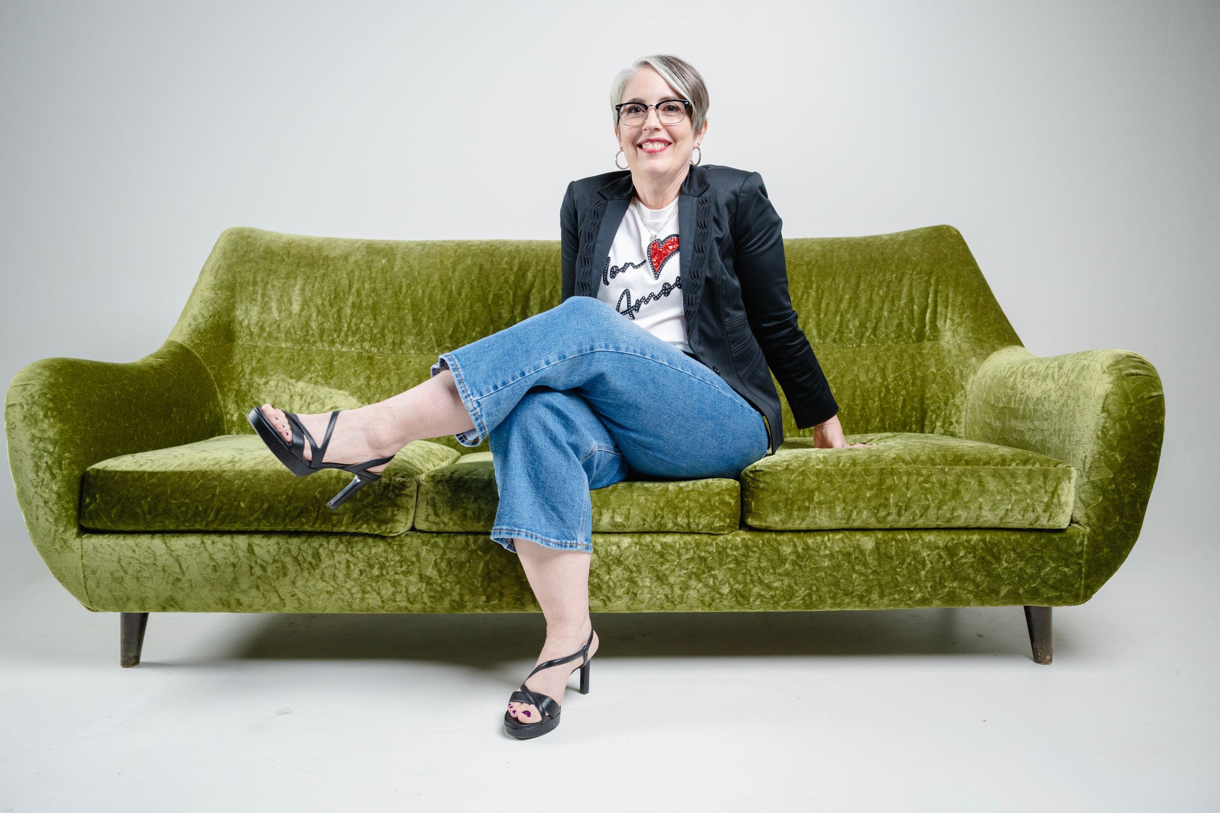 Someone relaxes casually on a retro green velvet sofa wearing a black blazer and jeans in a bright studio setting.