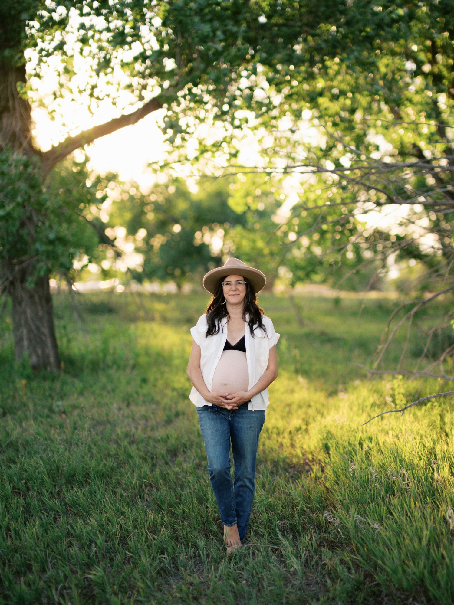Expecting mother in a sunlit meadow wearing a straw hat, white shirt and jeans during golden hour.