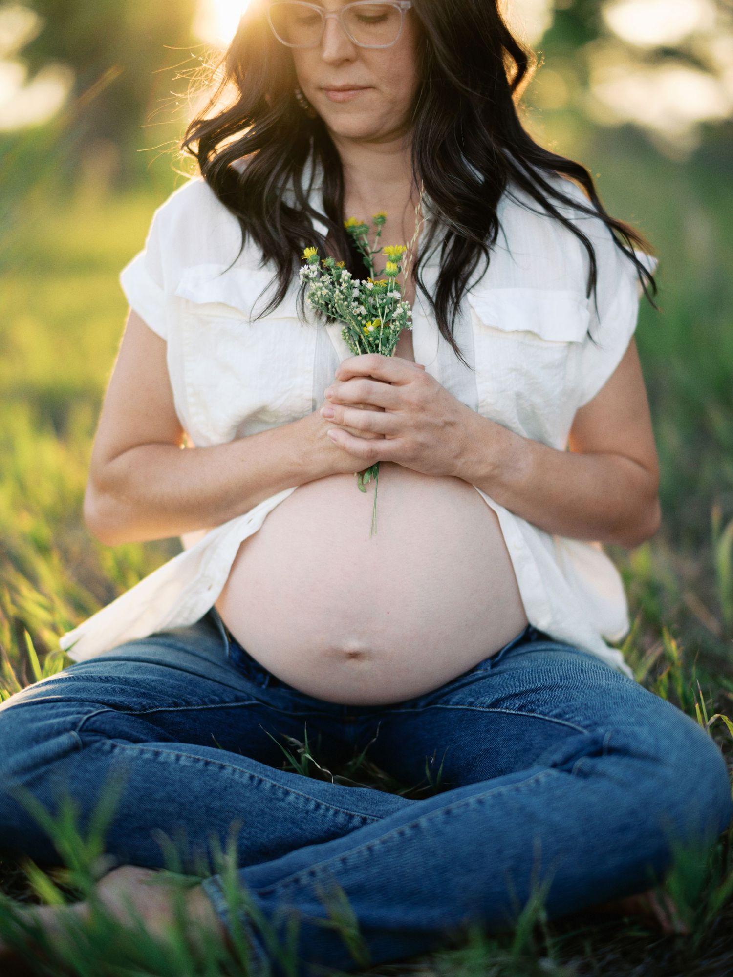 Expectant mother holding wildflowers while sitting in grass during sunset maternity photoshoot.