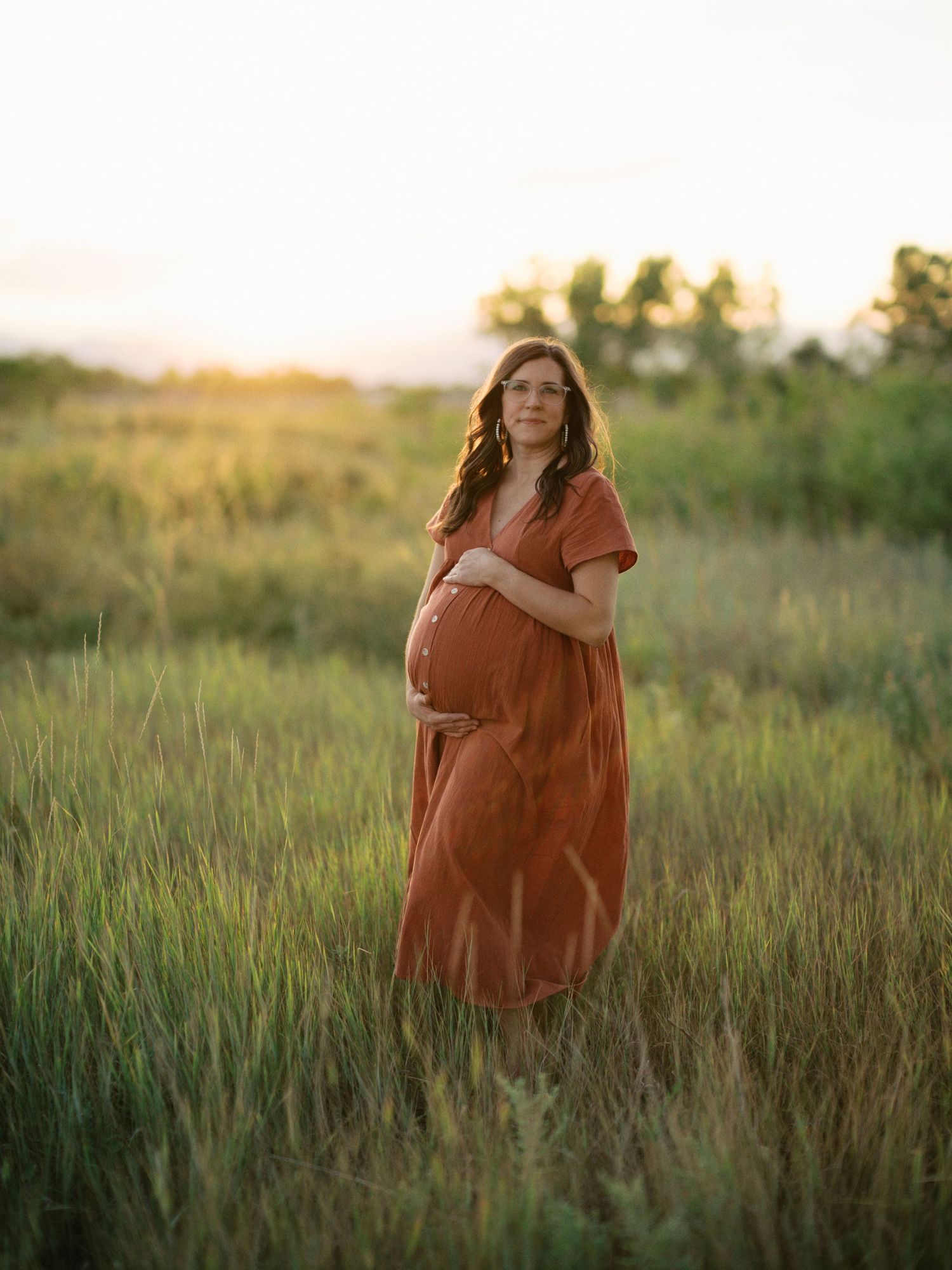 Pregnant woman in flowing rust-colored dress standing in tall grass during sunset.