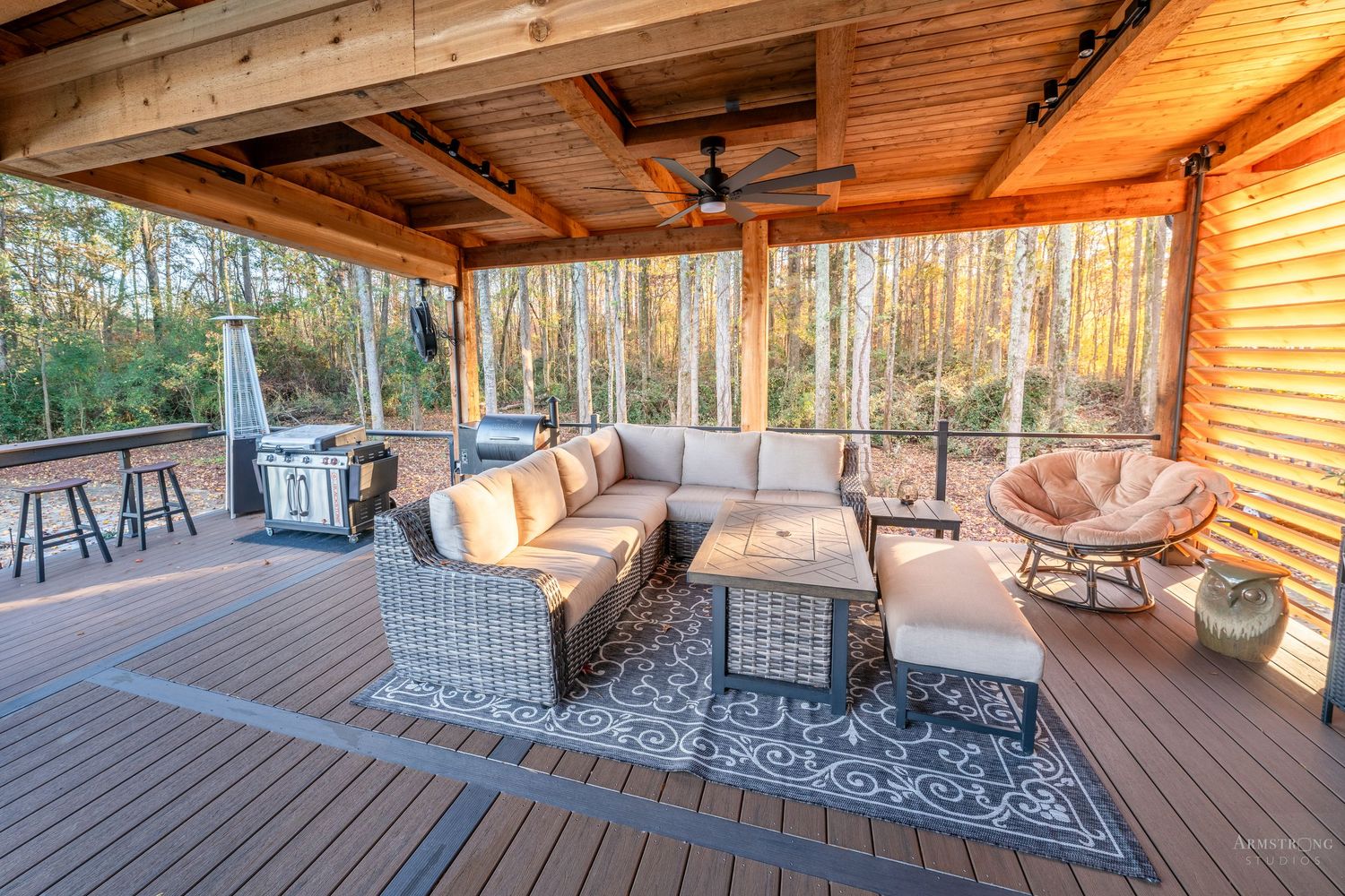 Cozy covered deck with wicker furniture, outdoor rug, and ceiling fan overlooking wooded backyard at sunset.