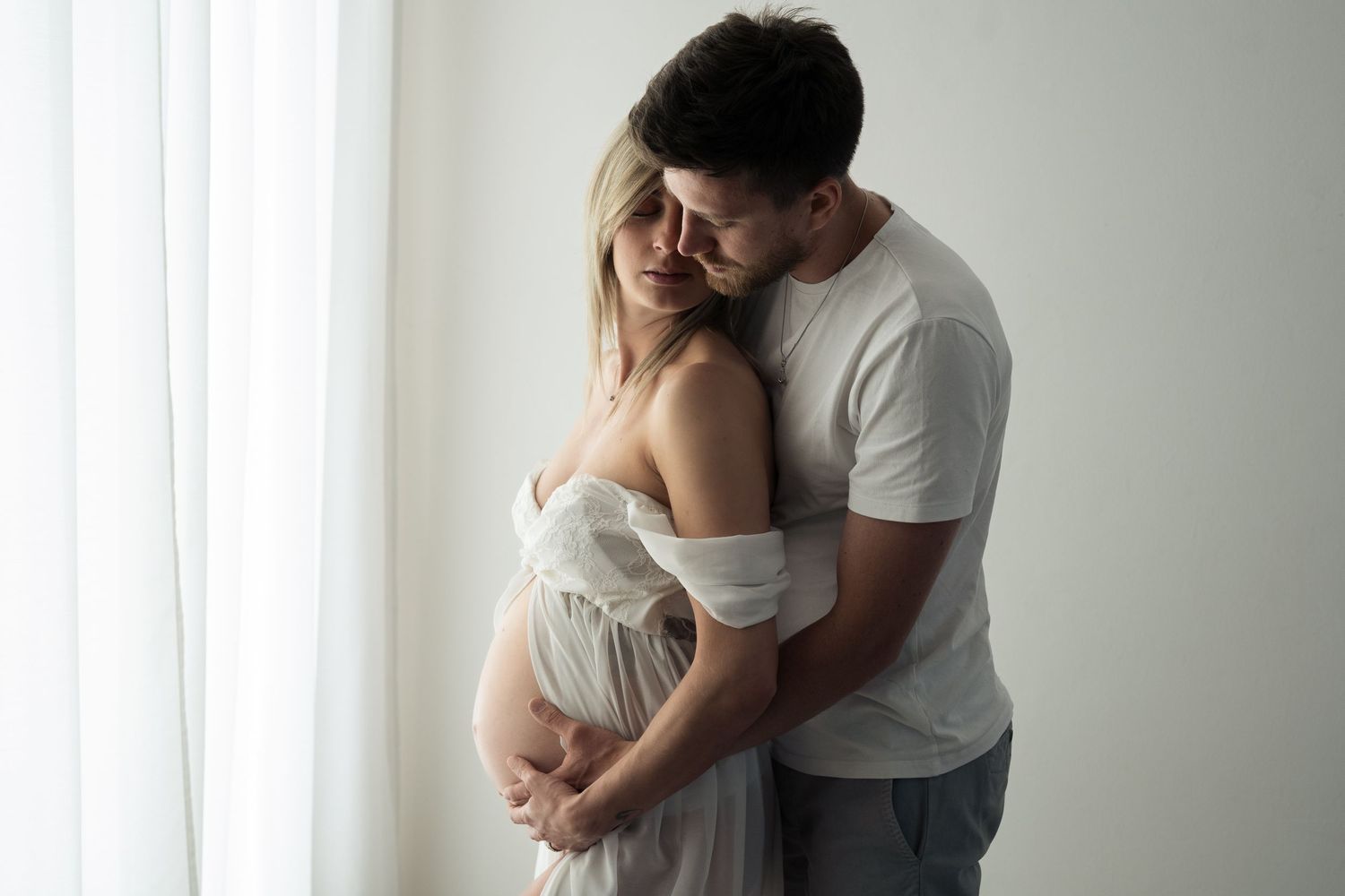 Donna in gravidanza abbracciata dal partner, entrambi con le mani sul pancione in un momento intimo e naturale