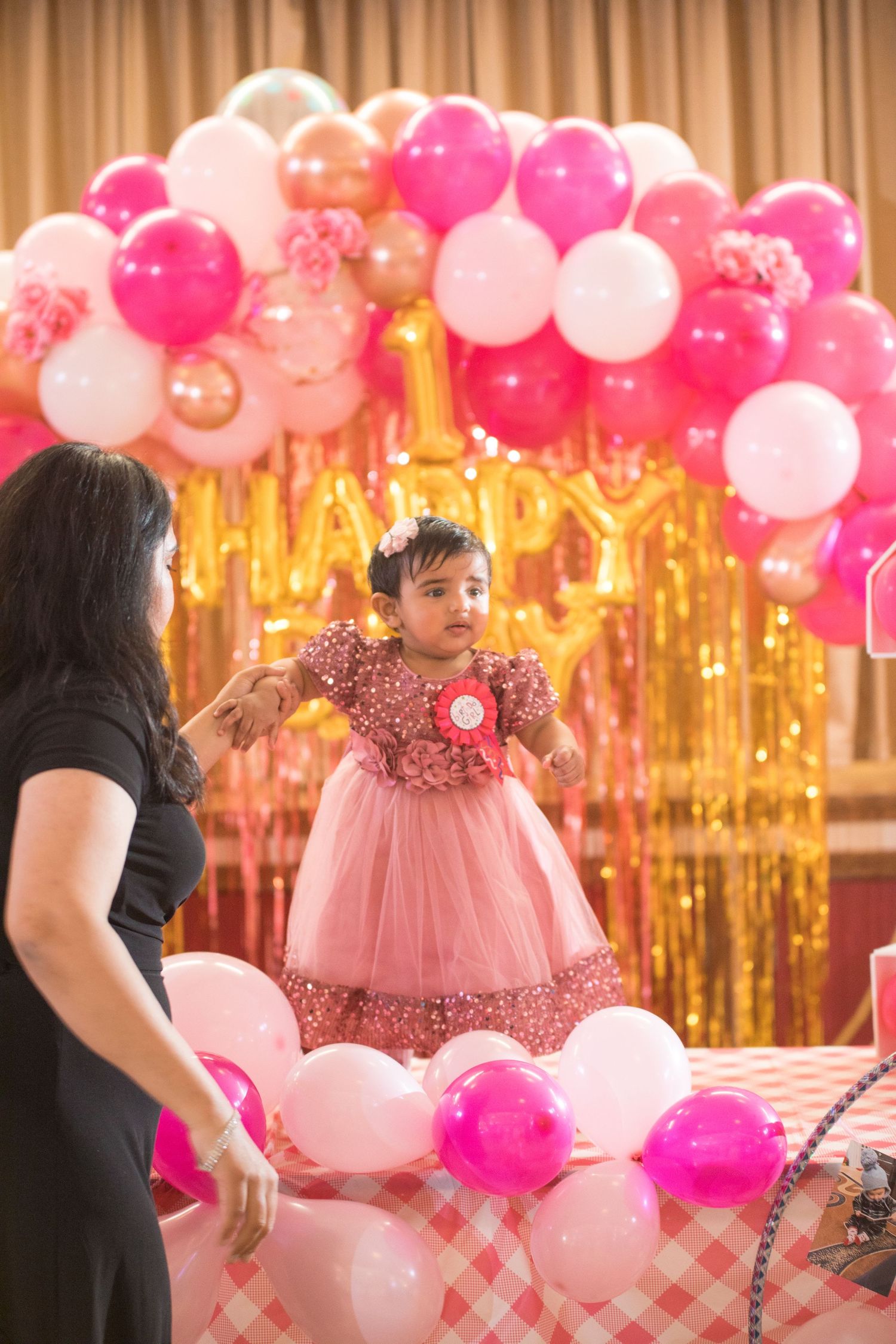 Child in pink dress standing on table near balloon arch at celebration.