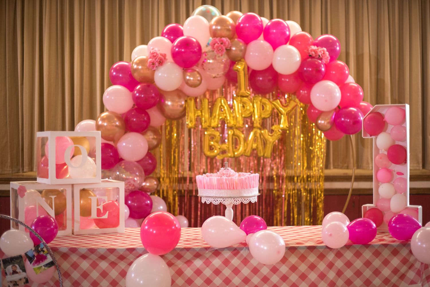Pink and white balloon arch with gold tinsel backdrop and 'Happy Birthday' sign.