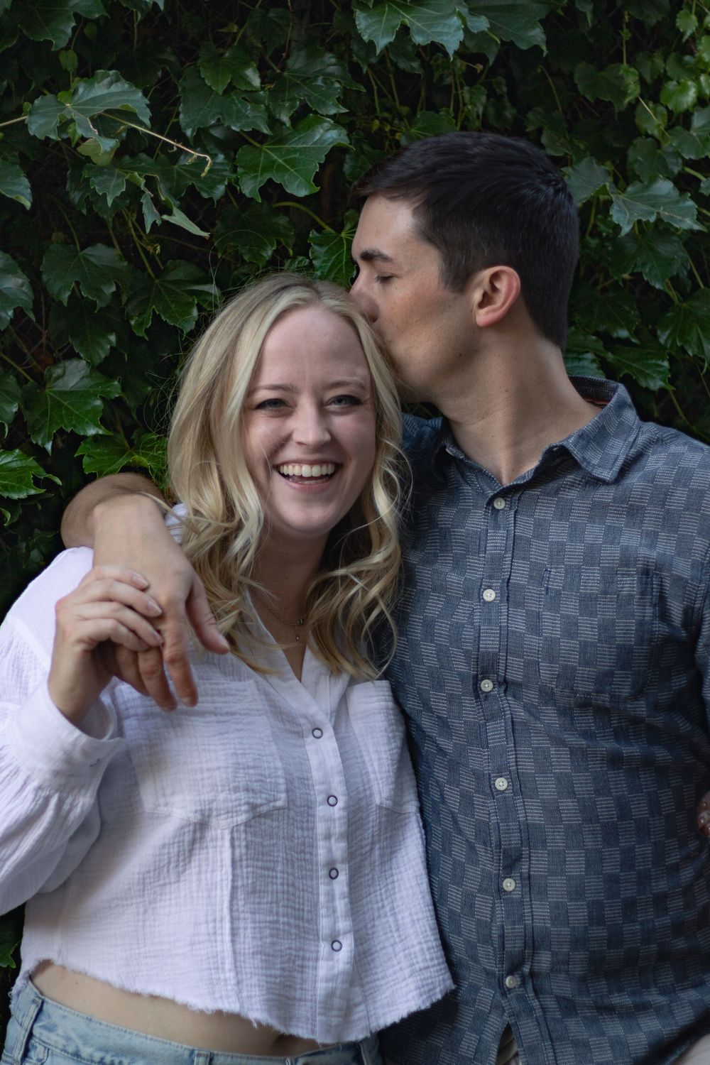 A couple laughs and embraces in front of a lush green wall.