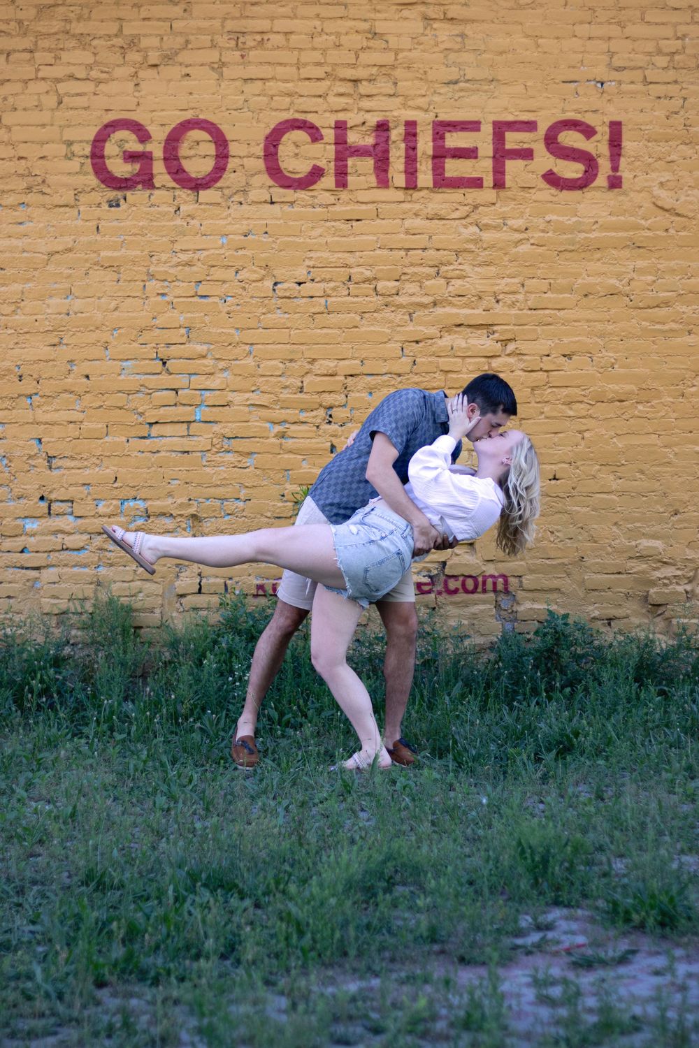 A couple shares a romantic dip pose in front of a brick wall with 'Go Chiefs' text.