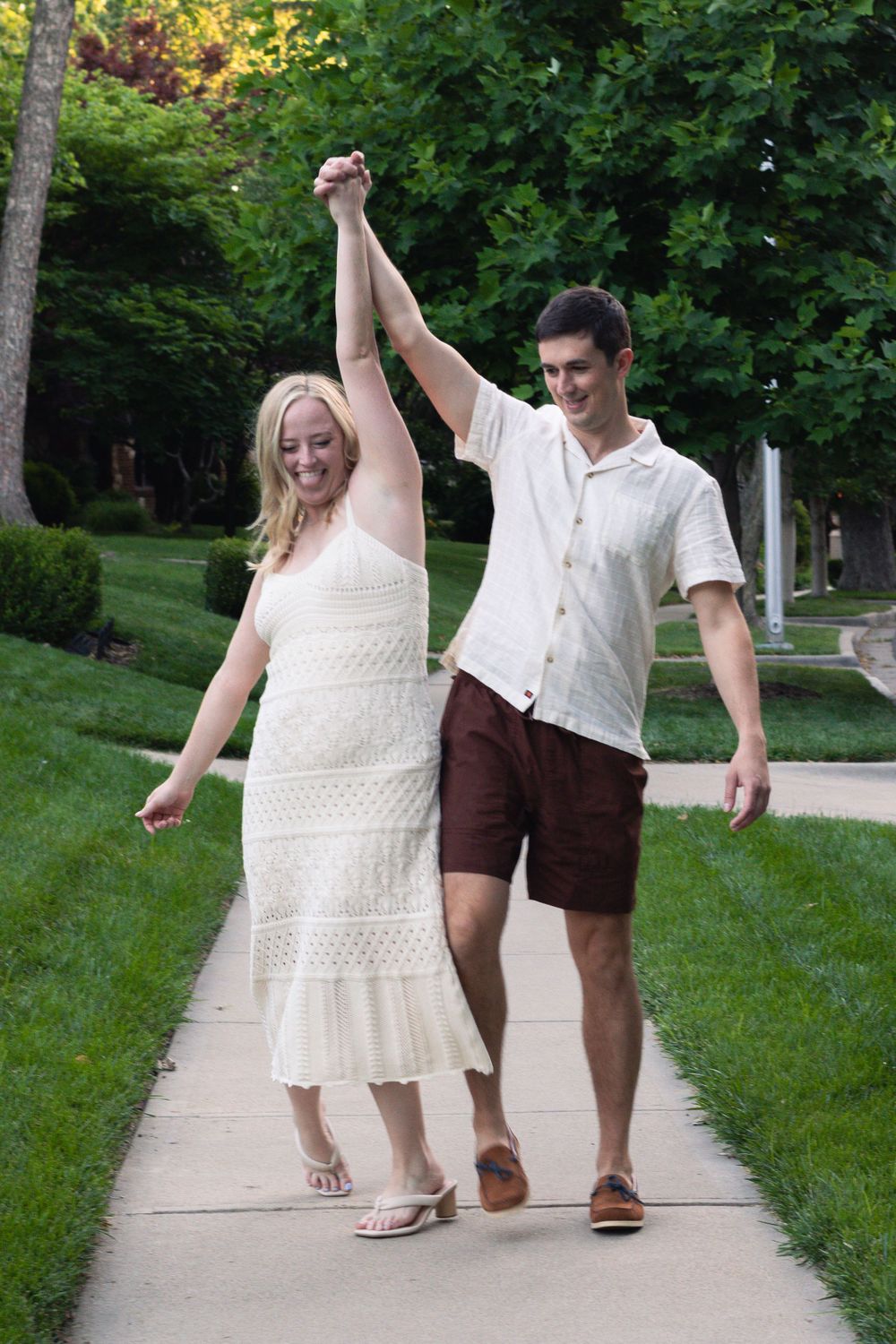 A couple walks together on a tree-lined path wearing summer outfits.