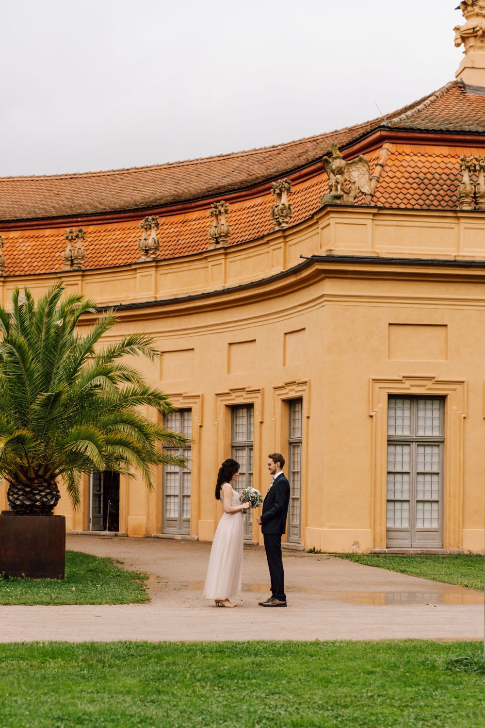 Wedding couple poses in front of a Spanish-style building with palm trees.