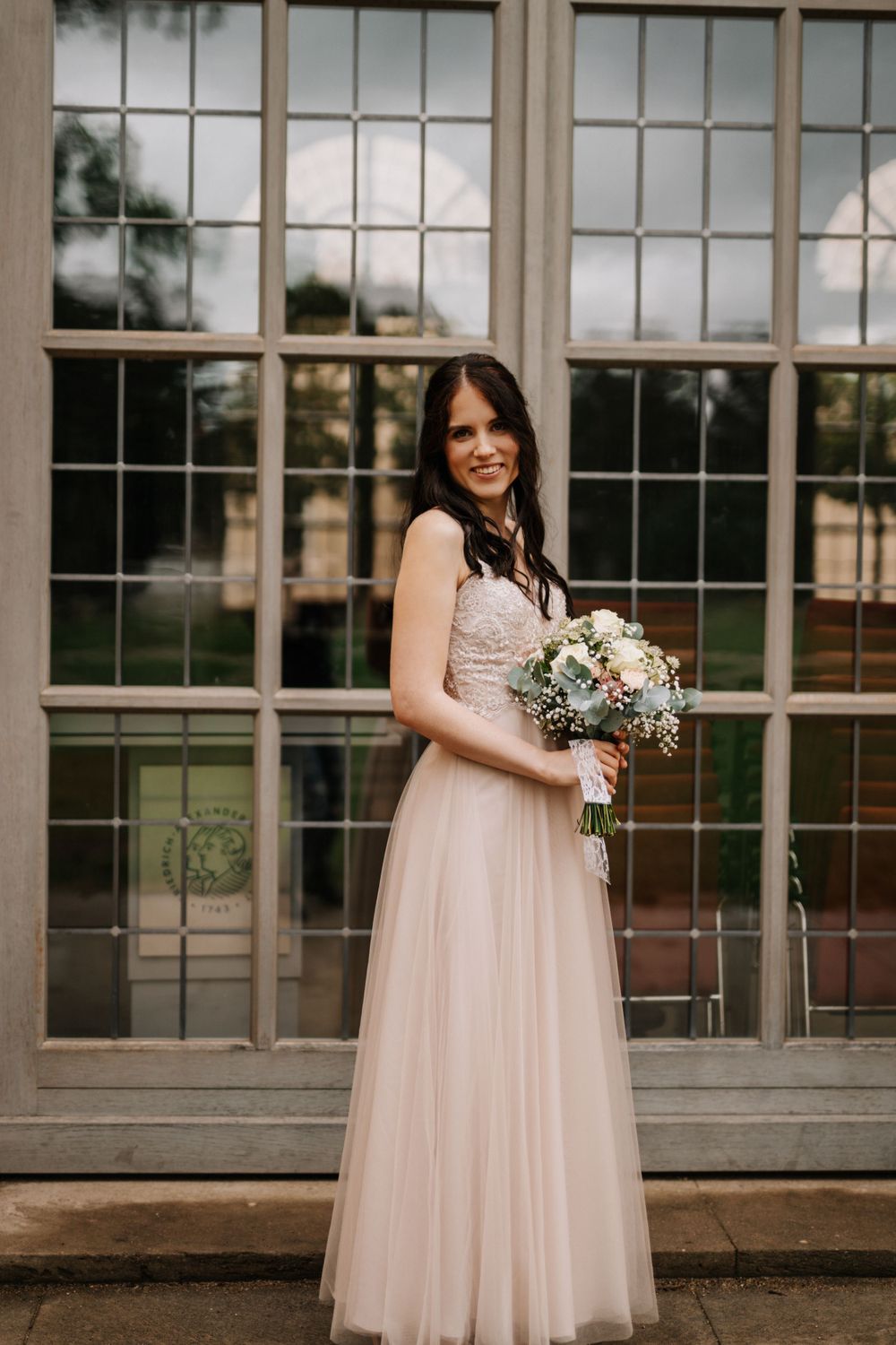 A bride in a blush gown poses joyfully in front of a historic building with large windows.