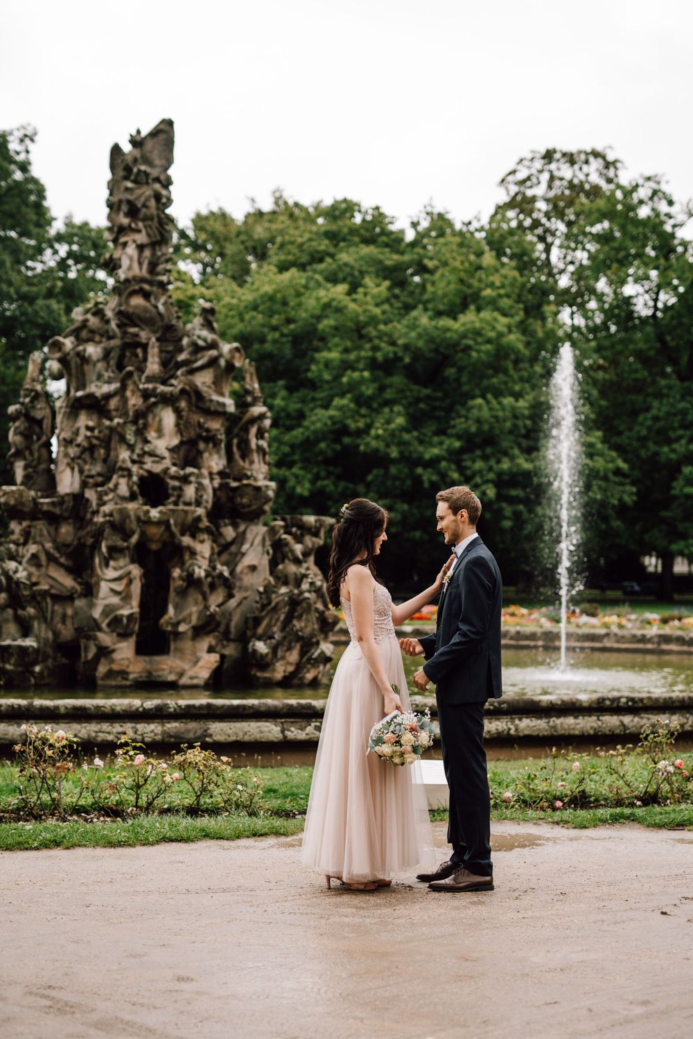 A couple shares an intimate moment in a garden with ornate stone sculptures and a fountain in the background.