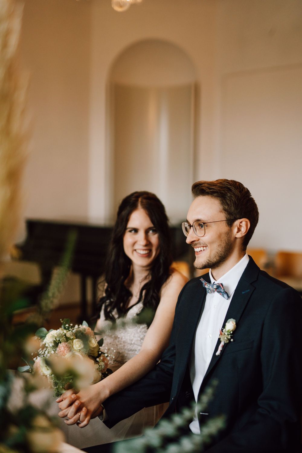 A couple sits together in formal attire surrounded by elegant decor.