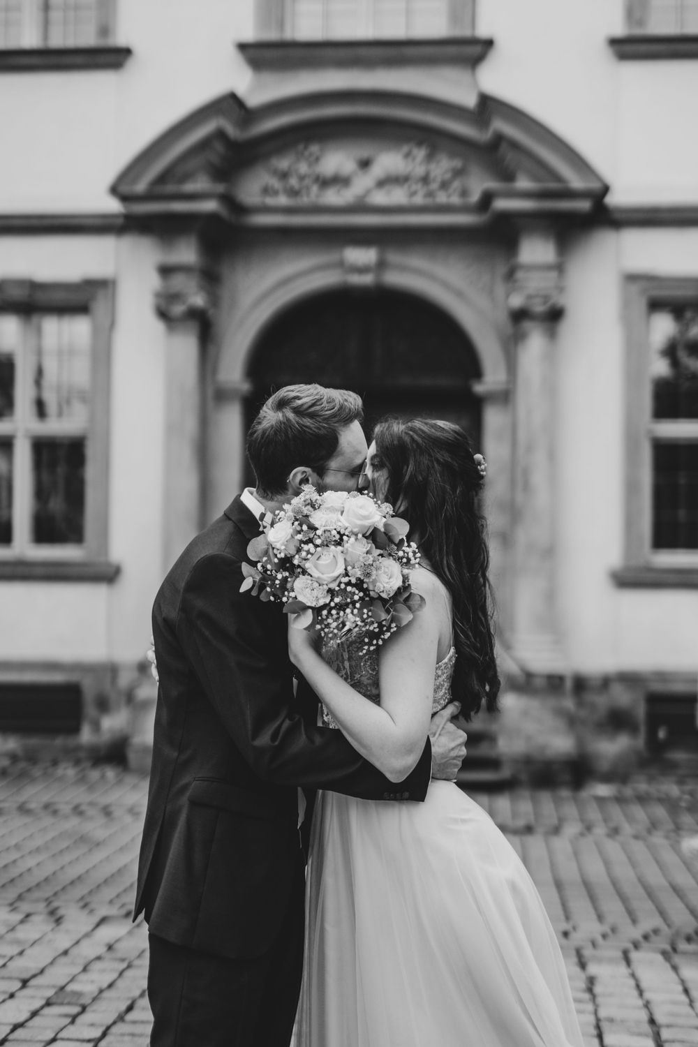 A couple embraces while holding a wedding bouquet outside a historic building.