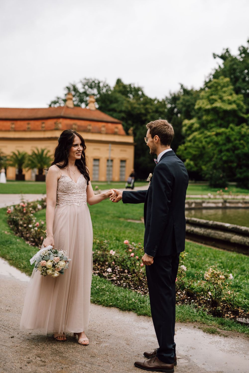 A couple in wedding attire walks along a garden path with blooming flowers and historic architecture.