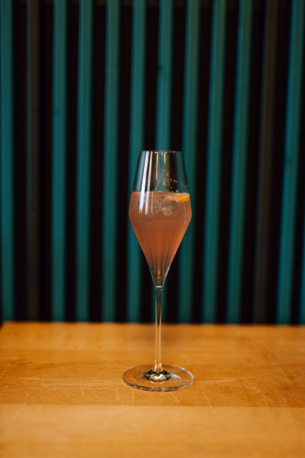 Single cocktail glass with orange-colored beverage on wooden table with teal curtain backdrop.