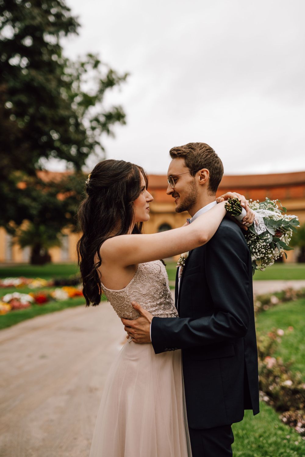 A romantic embrace captured in a garden setting with brick buildings and lush greenery.