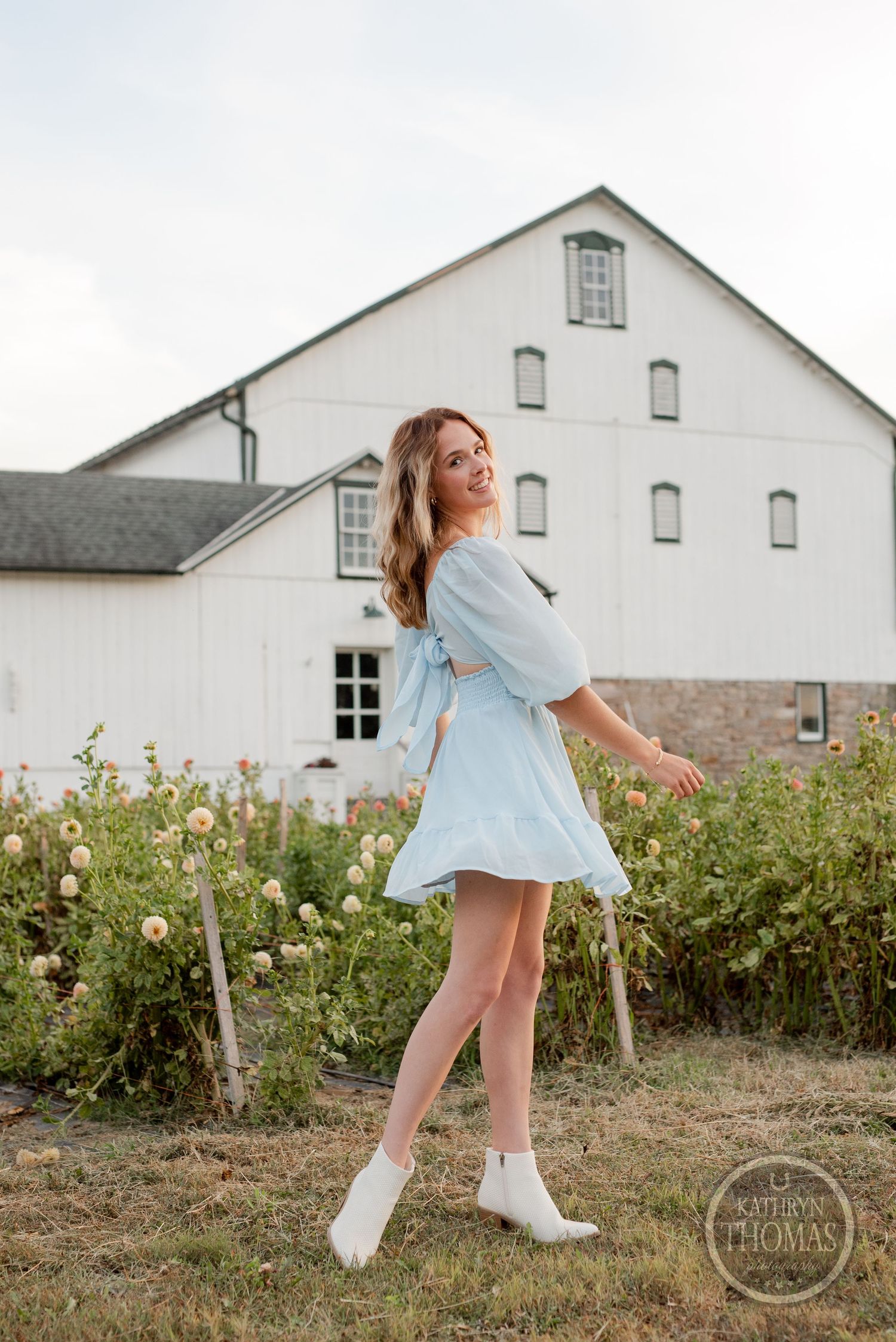 A light blue summer dress twirls against a rustic white barn backdrop with wildflowers in the foreground.