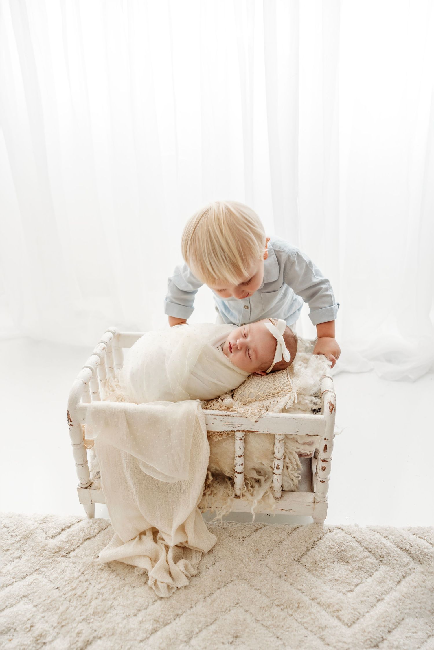 Baby in white with brother lying on rustic wooden crate with white lace backdrop in studio setting.Middle TN Newborn Photographer.