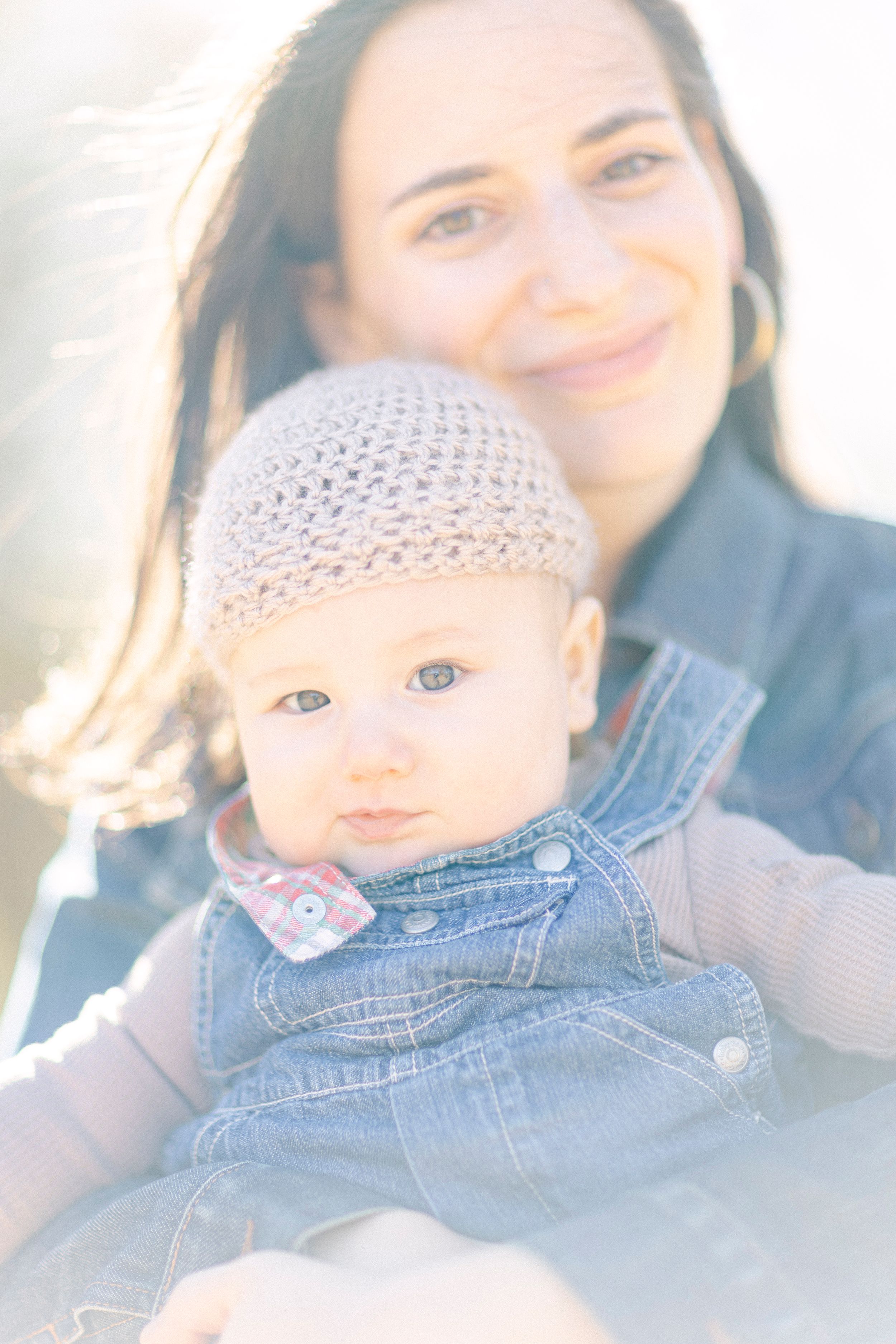 A baby in denim overalls and knit hat poses with parent in soft sunlit portrait.