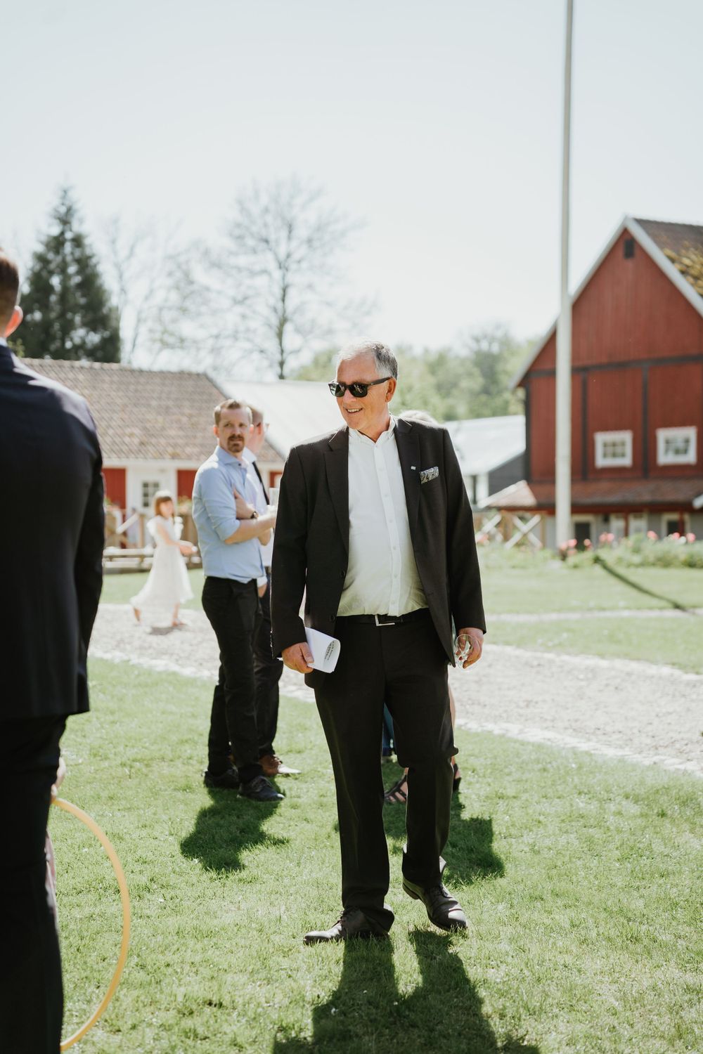 Guest in formal attire stands on grassy lawn with red barn buildings in background.