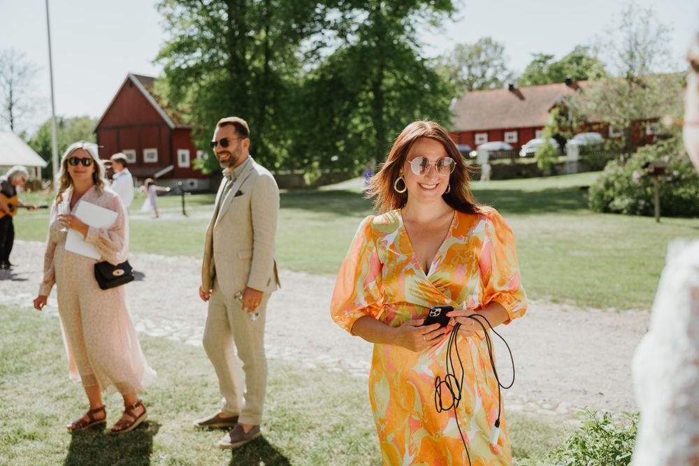 Wedding guests in bright summer attire gather outdoors near red barns.
